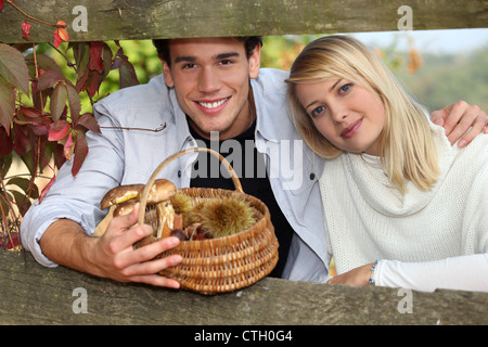 junges Paar hinter eine hölzerne Barriere im Herbst, der Mann hält einen Flechtwerk Korb mit Pilzen und Kastanien Stockfoto