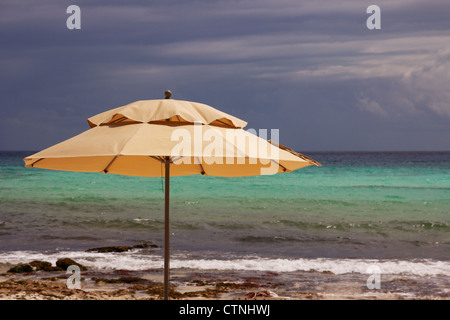 Tan Regenschirm vor dem Hintergrund der Brandung an einem natürlichen karibischen Strand und das türkisblaue Meer hinaus unter dunklen Wolken. Stockfoto