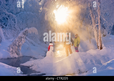 Ehemann Ehefrau stehen im frostigen Wald neben Schneemann Hintergrundbeleuchtung durch Sonnenstrahlen russische Jack Federn Park Yunan Alaska Winter Stockfoto