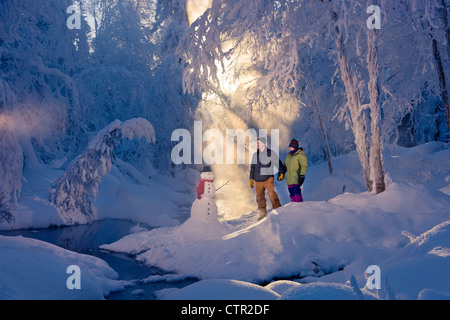 Ehemann Ehefrau stehen im frostigen Wald neben Schneemann Hintergrundbeleuchtung durch Sonnenstrahlen russische Jack Federn Park Yunan Alaska Winter Stockfoto