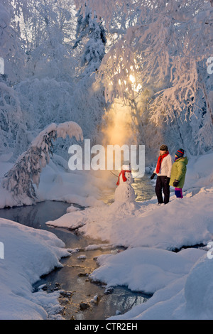 Zwei Frauen bewundern Schneemann tragen rote Schal schwarz Zylinderhut nächsten kleinen Bach in Raureif bedeckt Wald Russisch Jack Springs Stockfoto