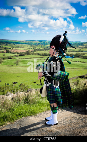 Kaukasischer Mann, der Dudelsäcke spielt, in Militärkostümen gekleidet, darunter ein traditioneller Kilt und eine Straußenfederhaube, in der Landschaft von Yorkshire. Stockfoto