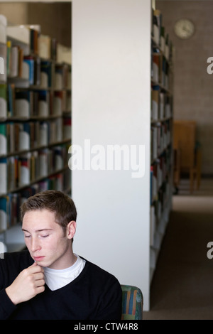 Ein junger Student an etwas denken oder lesen in der Bibliothek mit einer Wanduhr im Hintergrund sichtbar. Stockfoto