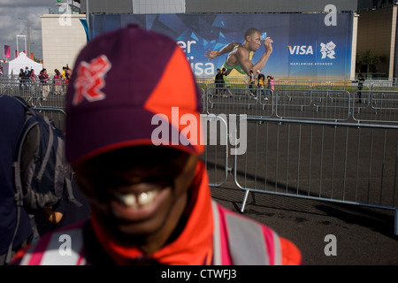 Ein Spiele-Hersteller freiwillig mit einem Goldzahn und Weitergabe Zuschauer in der Nähe von Usain Bolt Visa Plakat während der London 2012 Olympische Park während der Spiele. London 2012 Freiwillige nennt man "Spiele-Macher", wie sie dazu beitragen, um die Spiele geschehen zu machen. Bis zu 70.000 Spiele-Hersteller nehmen auf einer Vielzahl von Rollen über die Veranstaltungsorte: von Besuchern; für den Transport von Athleten; hinter den Kulissen in das Technik-Team aushelfen, um sicherzustellen, erhalten Sie die Ergebnisse so schnell und präzise wie möglich angezeigt. Spiele-Hersteller kommen aus den unterschiedlichsten Gemeinden und Hintergründen, aus in ganz Großbritannien Stockfoto