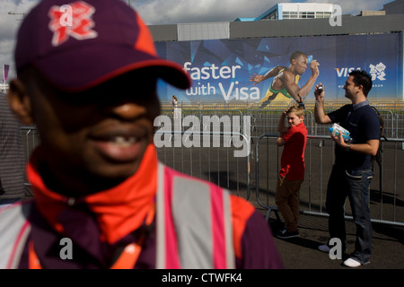 Ein Spiele-Hersteller freiwillig mit einem Goldzahn und Weitergabe Zuschauer in der Nähe von Usain Bolt Visa Plakat während der London 2012 Olympische Park während der Spiele. London 2012 Freiwillige nennt man "Spiele-Macher", wie sie dazu beitragen, um die Spiele geschehen zu machen. Bis zu 70.000 Spiele-Hersteller nehmen auf einer Vielzahl von Rollen über die Veranstaltungsorte: von Besuchern; für den Transport von Athleten; hinter den Kulissen in das Technik-Team aushelfen, um sicherzustellen, erhalten Sie die Ergebnisse so schnell und präzise wie möglich angezeigt. Spiele-Hersteller kommen aus den unterschiedlichsten Gemeinden und Hintergründen, aus in ganz Großbritannien Stockfoto