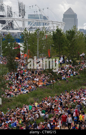 Massen der Zuschauer versammeln sich auf dem Rasen im Olympiapark, großen TV-Bildschirmen von live-Sport-Berichterstattung während der Olympischen Spiele 2012 in London zu sehen. Die Anpflanzung von 4.000 Bäume, 300.000 Feuchtgebietspflanzen und mehr als 150.000 Stauden sowie nektarreichen Wildblumen sorgen für eine farbenfrohe Kulisse für die Spiele. Dieses Land wurde umgestaltet, um eine 2,5 qkm sportliche Komplex, einmal Industriebetriebe und nun Austragungsort der acht Orte, darunter die Hauptarena Aquatics Centre und Velodrom sowie der Athleten Olympisches Dorf werden. Nach den Olympischen Spielen ist der Park als Queen Elizabeth Olympic Park bekannt. Stockfoto