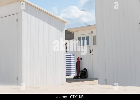 White Beach Huts stehend an einem weißen Sandstrand. Skateboard & Rucksack gegen eine Hütte & eine gestreifte Windschutz in der Ferne. Stockfoto