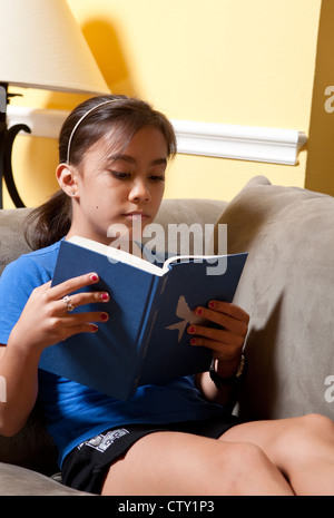 Zehn Jahre altes japanisch-amerikanische Mädchen liest einen Hardcover-Buch während Sie gemütlich auf dem Sofa zu Hause Stockfoto