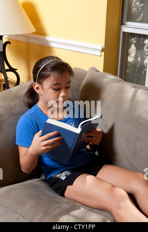 Zehn Jahre altes japanisch-amerikanische Mädchen liest einen Hardcover-Buch während Sie gemütlich auf dem Sofa zu Hause Stockfoto