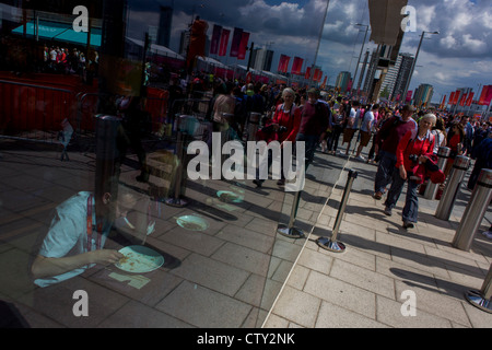 Gesehen durch das Fenster eines Fensters Westfield Shopping Mall, reinigt Olympic Angestellter seinen Teller während der Olympiade 2012 in London, die 30. Olympiade. Mit den Massen von ankommenden Zuschauer im Hintergrund reflektiert sehen wir die Banner der Spiele in London an einem brillanten sonnigen Tag in Stratford, im Osten Londons. Westfield liegt am Rande des Olympiaparks 2012, Europas größtes städtisches Einkaufszentrum bietet die Hauptzufahrt zum Olympiapark mit einem zentralen 'Straße' 75 % der Olympischen Besuchern den Zugang zu das Hauptstadion geben also Einzelhandelsflächen. Stockfoto