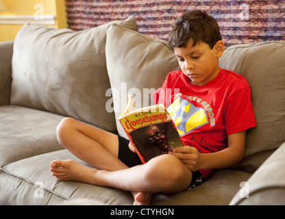 Der achtjährige mexikanisch-amerikanische Junge liest Softcover-Buch, während er sich zu Hause auf dem Sofa entspannt Stockfoto