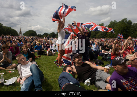 Britische Zuschauer feiern eine Goldmedaille gewinnen durch Team GB Triathlet Alistair Brownlee beim Triathlon, während der Olympiade 2012 in London, die 30. Olympiade im Hyde Park statt. Mädchen-Sport-Fans Welle ihrer Union Jack-Flagge über ihren Köpfen, als Tausende zu einen riesigen TV-Bildschirm mit dem Albert Memorial in der Ferne sehen. Brownlee kam zum ersten Mal gefolgt von Spaniens Javier Gomez dann Jonathan Brownlee (Bruder des Gewinners). (Weitere Beschriftungen in Beschreibung)... Stockfoto