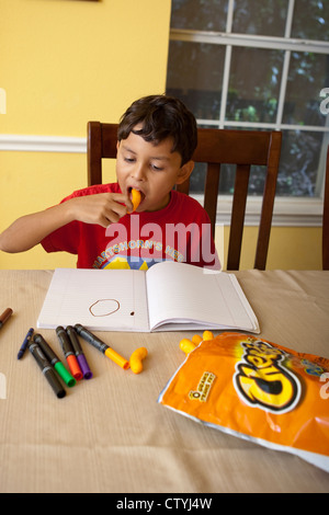 Ein achtjähriger mexikanisch-amerikanischer Knabberjause auf Junk Food, Cheetos, während er zu Hause mit Markern zeichnet. Stockfoto