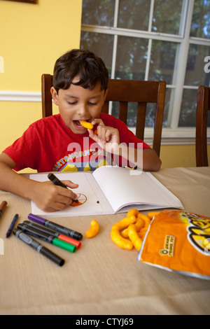 Ein achtjähriger mexikanisch-amerikanischer Knabberjause auf Junk Food, Cheetos, während er zu Hause mit Markern zeichnet. Stockfoto