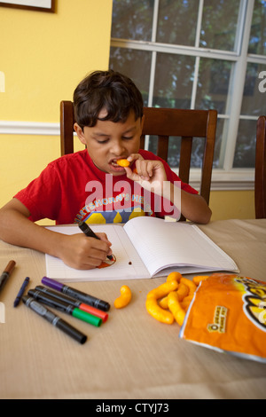 Ein achtjähriger mexikanisch-amerikanischer Knabberjause auf Junk Food, Cheetos, während er zu Hause mit Markern zeichnet. Stockfoto