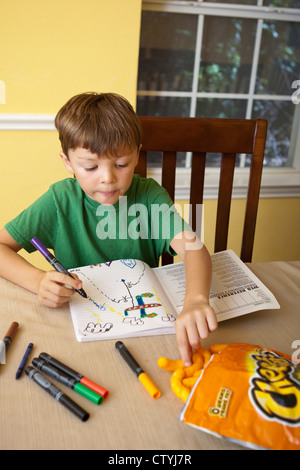 Ein siebenjähriger mexikanisch-amerikanischer Knabberjause auf Junk Food, Cheetos, während er zu Hause mit Markern zeichnet. Stockfoto