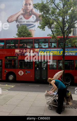 Eine Mutter füttert ihr Baby in der Nähe von der inspirierenden Bild von Team GB Goldmedaillengewinner Siebenkämpferin Jessica Ennis die das äußere des Adidas Store in der Londoner Oxford Street, während die Olympischen Spiele 2012 in London ziert. Die Anzeige ist für Schuhe Sportmarke Adidas und ihre "Nehmen The Stage"-Kampagne, die sichtbar ist, in ganz Großbritannien und Briten, die diese Athleten gejubelt haben, die gewonnen Medaillen in Zahlen seit 100 Jahren nicht mehr gesehen haben. Stockfoto