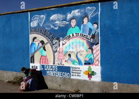 Die gebürtige Frau sitzt auf dem Bürgersteig neben einem Wandgemälde, das Teil einer Kampagne zur Reduzierung von häuslicher Gewalt gegen Frauen ist, El Alto, Bolivien Stockfoto