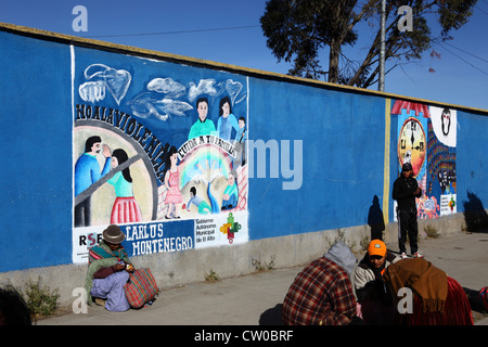 Die gebürtige Frau sitzt auf dem Bürgersteig neben einem Wandgemälde, das Teil einer Kampagne zur Reduzierung von häuslicher Gewalt gegen Frauen ist, El Alto, Bolivien Stockfoto