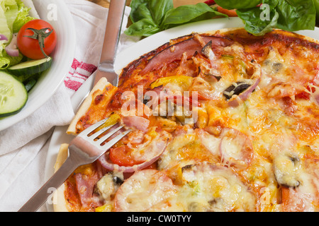 Gebackene Pizza mit Schinken, Gemüse, Basilikum und frischen Salat, Stillleben als Nahaufnahme mit Messer und Gabel für italienisches Essen Stockfoto