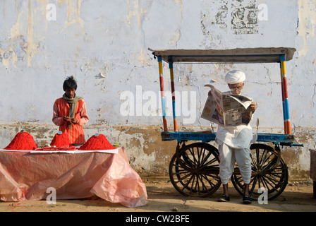 Älterer Mann lesen Zeitung. Am Wegesrand Anbieter verkaufen Farbe Farbstoffe neben ihm - Pushkar, Rajasthan Stockfoto