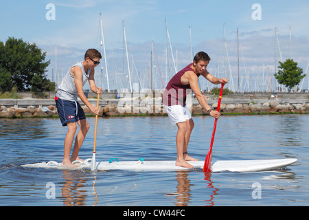 Zwei Teenager Spaß Stand up Paddeln auf einem windsurfboard an einem sonnigen Sommertag am Strand Rungsted Hafen, Dänemark. Paddleboarders. Stockfoto