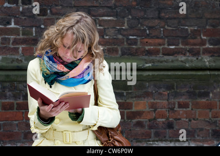 junge Frau in einem Mantel sitzt vor einer roten Backsteinmauer, ein Buch zu lesen Stockfoto