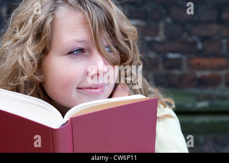 junge Frau in einem Mantel sitzt vor einer roten Backsteinmauer aus einem offenen Buch nachschlagen Stockfoto