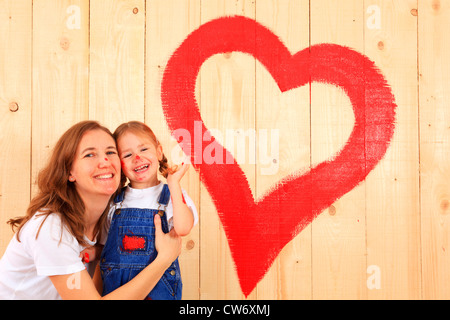 kleines Mädchen hat ein Herz auf einer Holzwand mit roter Farbe gemalt, Mutter nahm sie in die Arme mit einem Lächeln Stockfoto