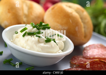 Frische Sahne Schmelzkäse in Schälchen mit Schnittlauch obendrauf mit Salami-Scheiben und Brötchen auf Platte Stockfoto