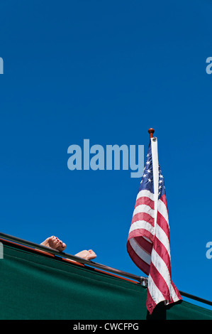 Eine Frau die Füße mit lackierten Zehennägeln ruhen auf dem Geländer eines Bootes neben einer amerikanischen Flagge vor einem blauen Himmelshintergrund. Stockfoto