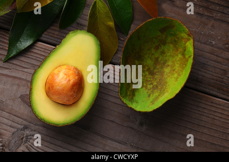 Eine Avocado halbieren auf eine Holzoberfläche mit Blättern. Eine Hälfte zeigt die Samen während der anderen ausgehöhlt. Stockfoto