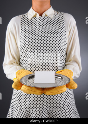 Nahaufnahme einer Hausfrau in eine Schürze und Backofen Handschuhe halten ein Tablett mit Tischkarte Plank. Stockfoto
