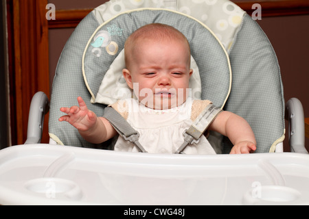 Ein 6 Monate altes Baby Mädchen, in einem Hochstuhl sitzen schreit während Sie darauf warten, gefüttert zu werden Stockfoto