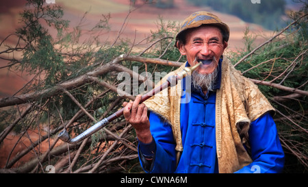 Ein lächelnder alter asiatischer Mann, der Pfeife raucht und auf seinem Rücken Äste trägt Stockfoto