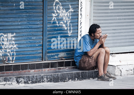 Mann Beleuchtung Zigarettenanzünder saßen draußen ein Shop in Bangkok. street scene von jemand nach unten und heraus in Bangkok. düsteren Hintergrund der Graffiti auf store Fensterläden Stockfoto