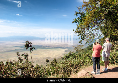 Ngorongoro-Krater Besucher des Lookout Tansania / NGORONGORO CONSERVATION AREA, Tansania — zwei Besucher fotografieren den weitläufigen Boden des Ngorongoro-Kraters von einem bewaldeten Aussichtspunkt entlang des Randes. Das sichtbare Becken darunter ist die größte intakte und unüberflutete vulkanische Caldera der Welt. Die steilen Mauern entstanden, als vor etwa zweieinhalb Millionen Jahren ein massiver Vulkan einbrach, bilden eine natürliche Einfriedung, die eine der dichtesten Konzentrationen Afrikas an Wildtieren das ganze Jahr über beherbergt. Stockfoto