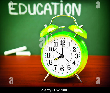 Foto von Alarm clock auf hölzernen Schreibtisch im Klassenzimmer, Handschrift Wort 'Bildung' am grünen Tafel, Herbstsaison, gelbe Uhr Stockfoto