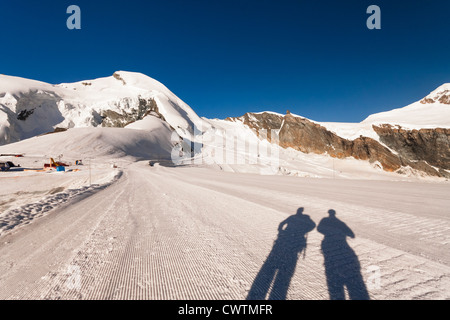 Die Schatten der beiden Bergsteiger auf der Piste Mittel Allalin in Saas Fee der Schweiz mit Allalinhorn in der Ferne. Stockfoto