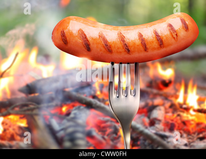 Wurst auf einer Gabel. Im Hintergrund ein Lagerfeuer im Wald. Stockfoto