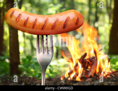 Wurst auf einer Gabel. Im Hintergrund ein Lagerfeuer im Wald. Stockfoto