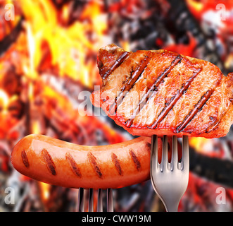 Wurst und Steak auf einer Gabel. Im Hintergrund ein Lagerfeuer im Wald. Stockfoto