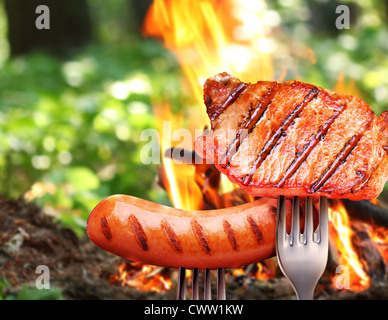 Wurst und Steak auf einer Gabel. Im Hintergrund ein Lagerfeuer im Wald. Stockfoto