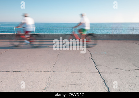 Zwei Personen auf gemieteten Fahrrädern am Meer. Bewegungsunschärfe. Eine Menge textfreiraum im Vordergrund und Meer und Himmel im Hintergrund. Stockfoto