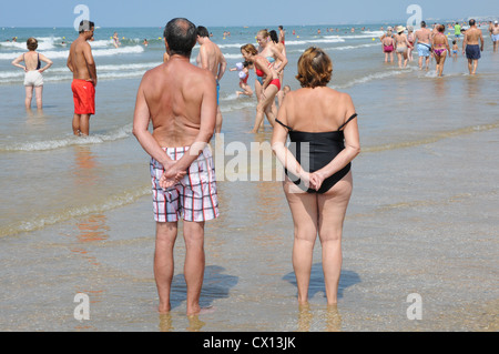 Rückseite älteres Paar, 60, stehend mit Hände hinter Rücken, beobachten andere Urlauber Sandstrand entlang schlendern. Stockfoto