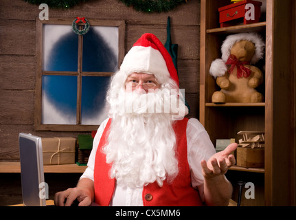 Santa Claus in seiner Werkstatt mit Laptop und umgeben von Spielzeug und Geschenke. Stockfoto