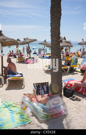 eine Frau liest eine Zeitschrift am Strand von Alcudia Mallorca unter den Sonnenliegen unter einer Palme Stockfoto