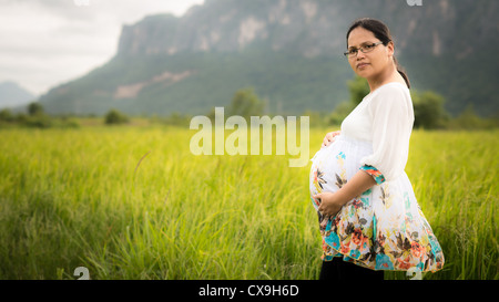 Schöne asiatische Frau trägt Brille und liebevoll hält ihren schwangeren Bauch in einem neu gepflanzten Reisfeld Stockfoto
