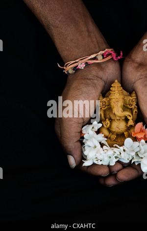 Hinduistische Elefantengott. Indische mans Hand mit Lord Ganesha Statue und Jasminblüten vor schwarzem Hintergrund Stockfoto