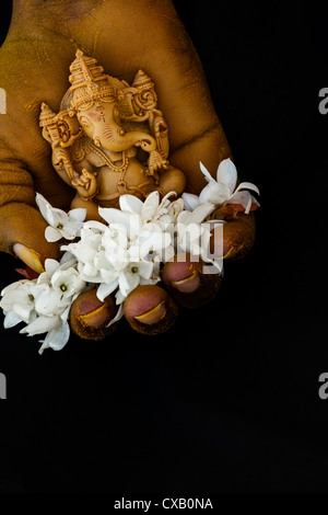 Hinduistische Elefantengott. Kurkuma bedeckt indischen mans Hand mit Lord Ganesha Statue und Jasmin Blüten auf schwarzem Hintergrund Stockfoto